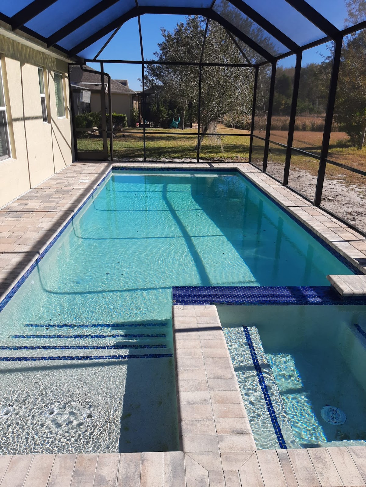 Rectangular residential swimming pool and spa inside a screened enclosure, with clear blue water, decorative tile, and light stone decking.