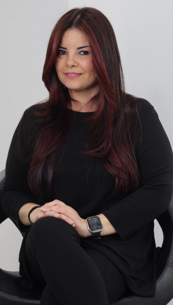 A woman with long, wavy, dark red hair is sitting in a chair, looking at the camera with a slight smile. She is wearing a black long-sleeved shirt and black pants. Her hands are clasped in her lap, and she is wearing a smartwatch on her left wrist. The background is a plain white wall.