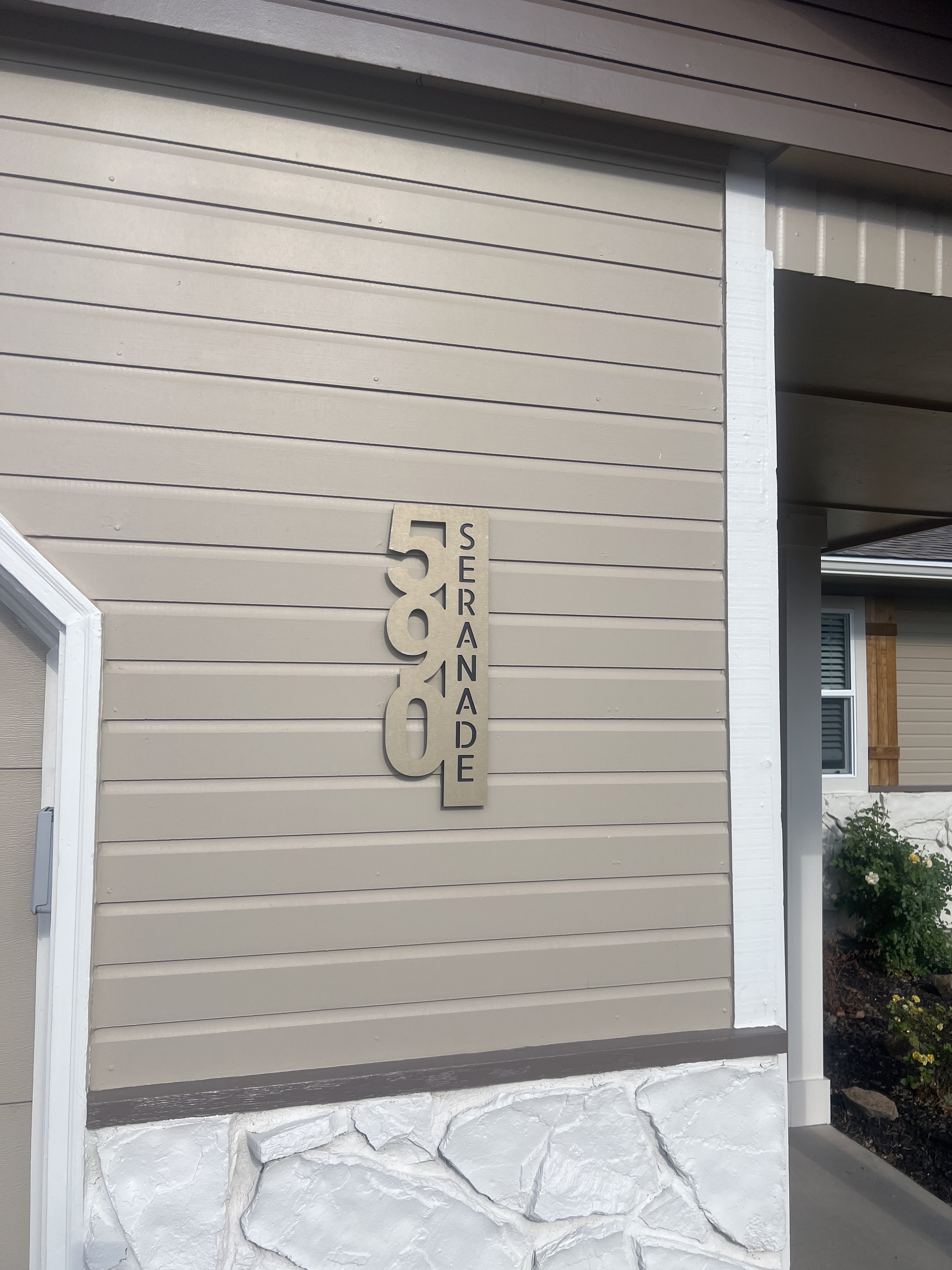 A beige vinyl-sided house exterior with a white trim around a doorway is visible. A modern, silver house number sign reads "588 Serenade." The sign is mounted vertically on the siding. A covered porch with a white column and dark ceiling is to the right. In the background, a window with blinds and wooden shutters is visible above a stone foundation. Landscaping with mulch and a flowering bush with yellow roses is present.