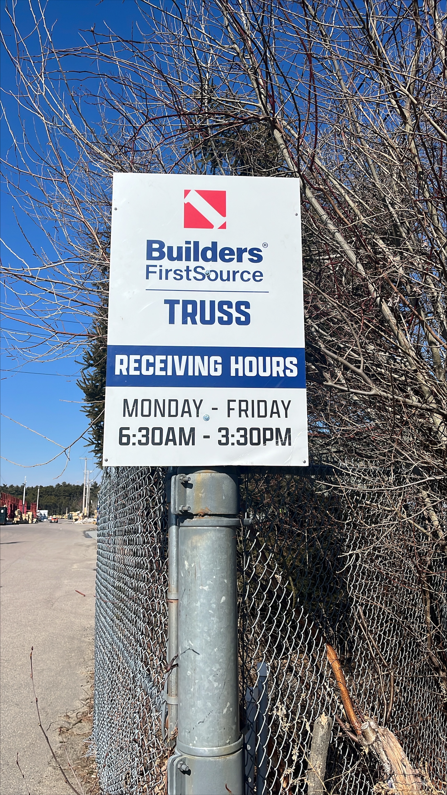 A close-up of a Builders FirstSource sign displaying truss receiving hours, indicating operational times from Monday to Friday, 6:30 AM – 3:30 PM. The sign is mounted on a metal post along a chain-link fence, with a construction yard in the background.