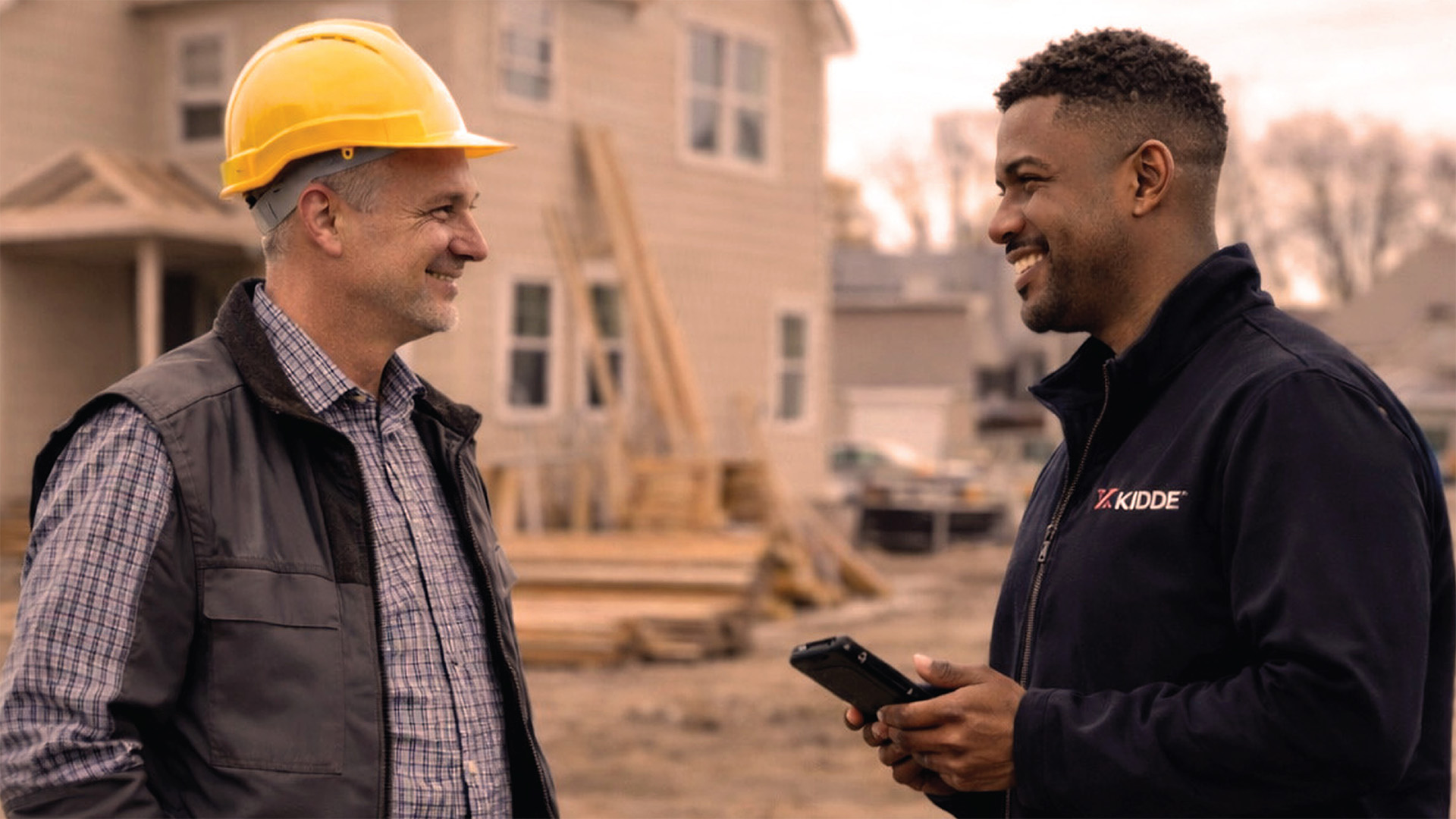 Two contractors are discussing a project, an in-progress home is in the background.