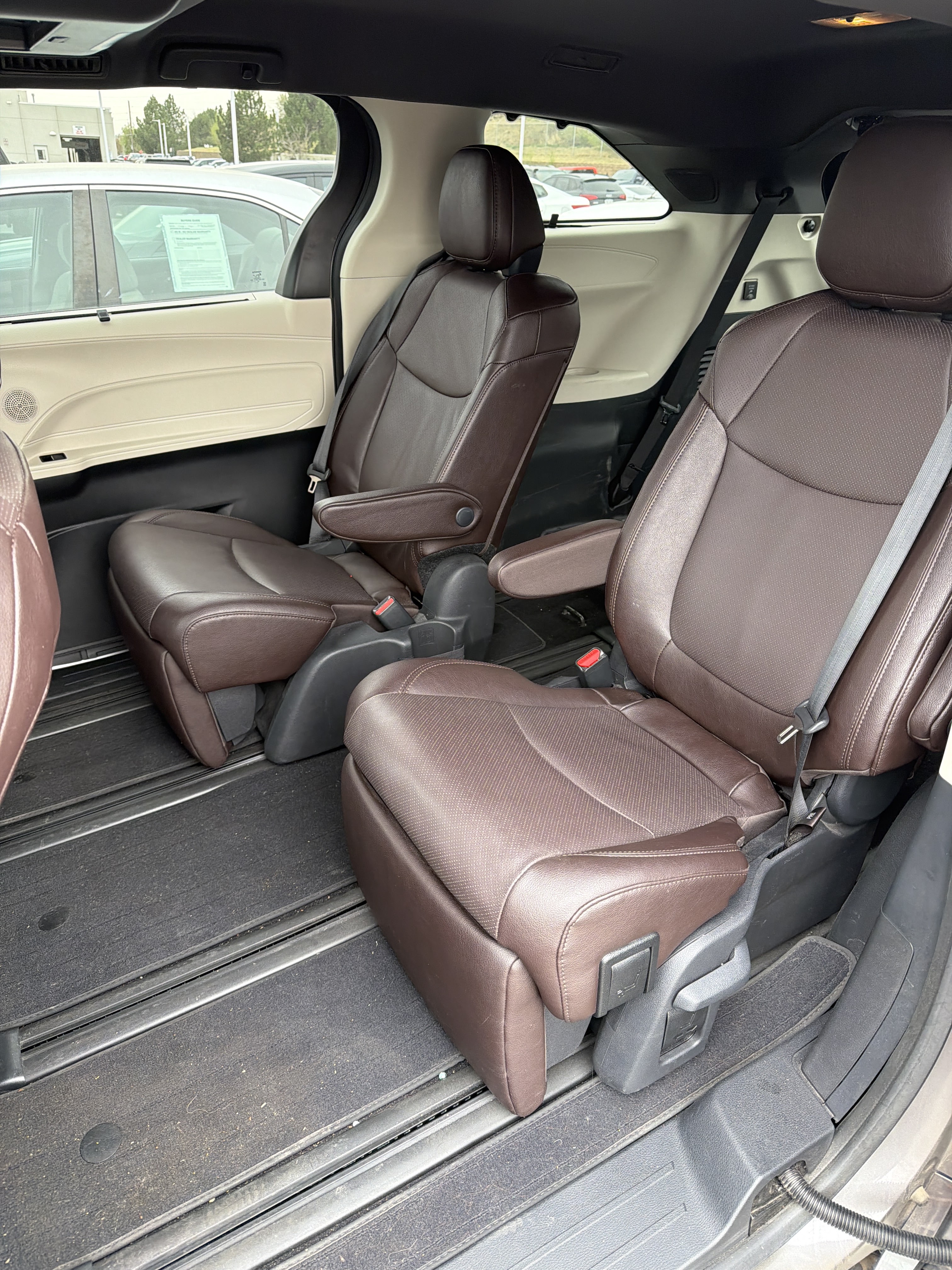 A close-up view of the interior of a spacious van, focusing on the brown leather captain's chairs. The seats have perforated inserts and adjustable armrests. The floor is covered in black carpet with visible silver rails for seat adjustment. In the background, through the windows, a car dealership lot with multiple vehicles and a building can be seen.