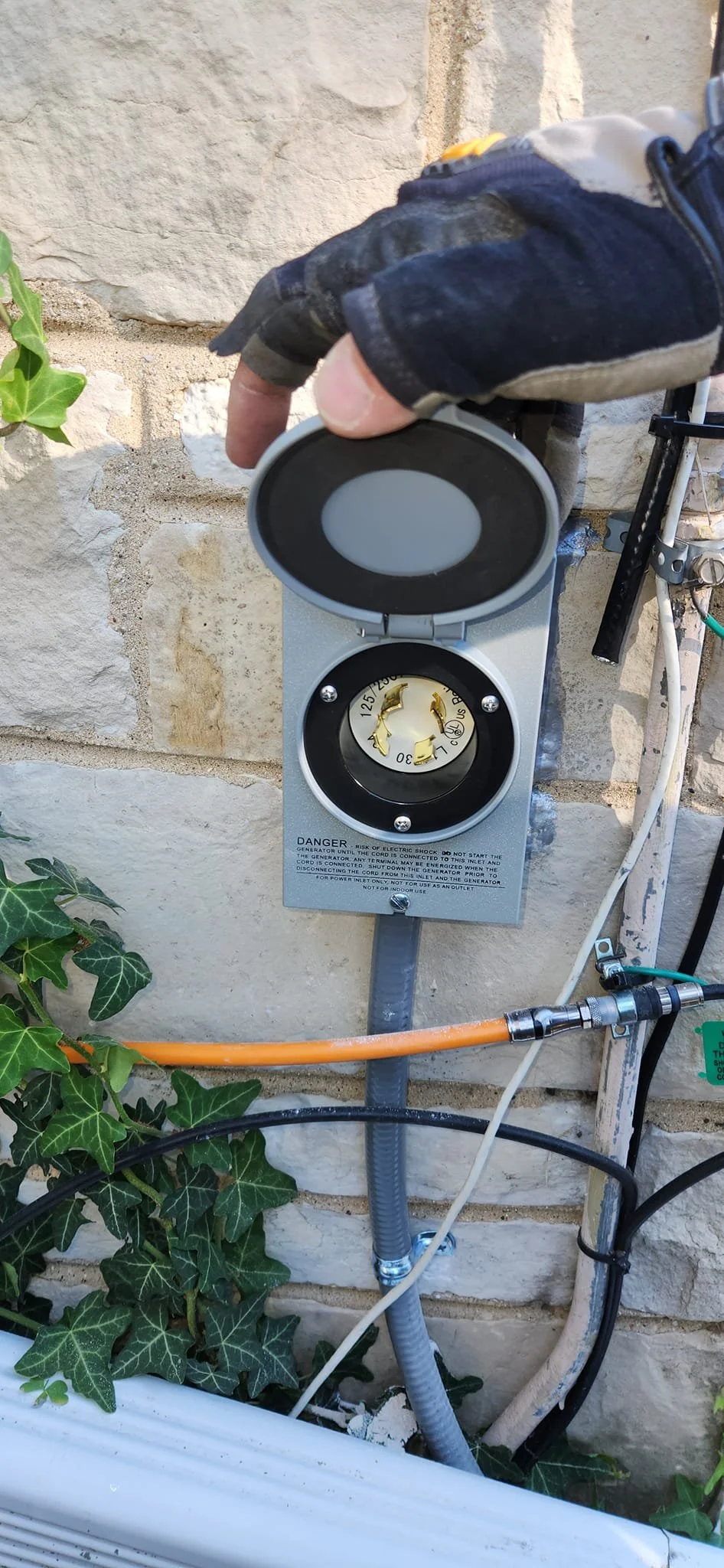 A hand wearing a work glove is opening a gray electrical inlet box mounted on a stone wall. The inlet is labeled "DANGER" and "125/250V L30". Various electrical wires and conduits are visible around the inlet box, along with ivy growing on the wall.