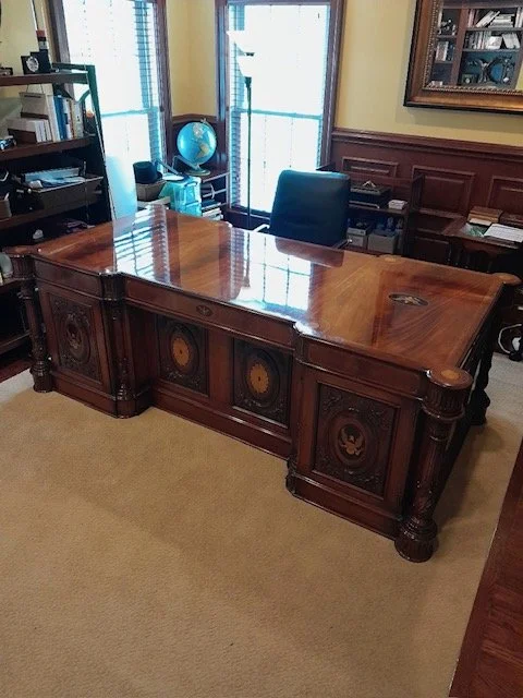 A large, ornate wooden desk is the focal point of this office scene. The desk is highly polished and reflects the light, showcasing intricate carvings and detailing. Behind the desk, a black leather office chair is visible, positioned in front of two large windows with blinds. To the left of the desk, a bookshelf is filled with books and various items, including a globe. The walls are paneled with wood, and a framed picture hangs on the right wall. The floor is covered with a light brown carpet.