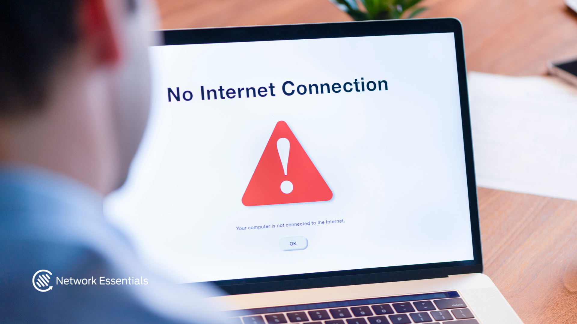 A person, seen from behind, looks at a laptop screen displaying a "No Internet Connection" error message. The error message includes a red triangular warning sign with an exclamation mark and the text "Your computer is not connected to the Internet.", along with an "OK" button. A logo for "Network Essentials" is visible in the bottom left corner. The scene appears to be a desk with a wooden surface.