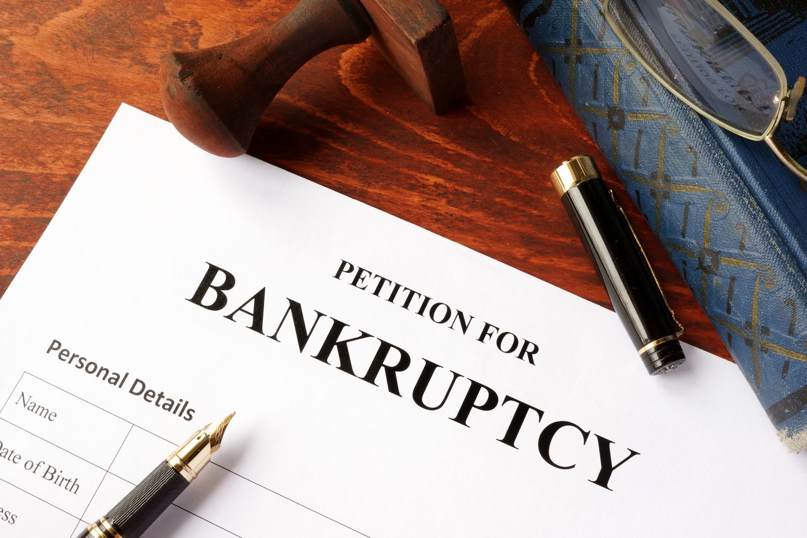 A "Petition for Bankruptcy" form sits on a polished wooden desk beside a fountain pen, its black capped lid, a wooden rubber stamp, eyeglasses, and an antique blue book.