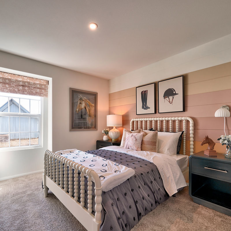 A well-decorated bedroom with a horse-themed design. The room features a white spindle bed with gray bedding and a blanket adorned with horseshoe patterns. Two framed artworks of equestrian items, a horse's head, and a rider's helmet hang on a horizontally striped wall in pastel shades of pink, peach, and beige. A bedside table with a lamp and a horse figurine complements the theme. A large window with a patterned Roman shade allows natural light into the room.