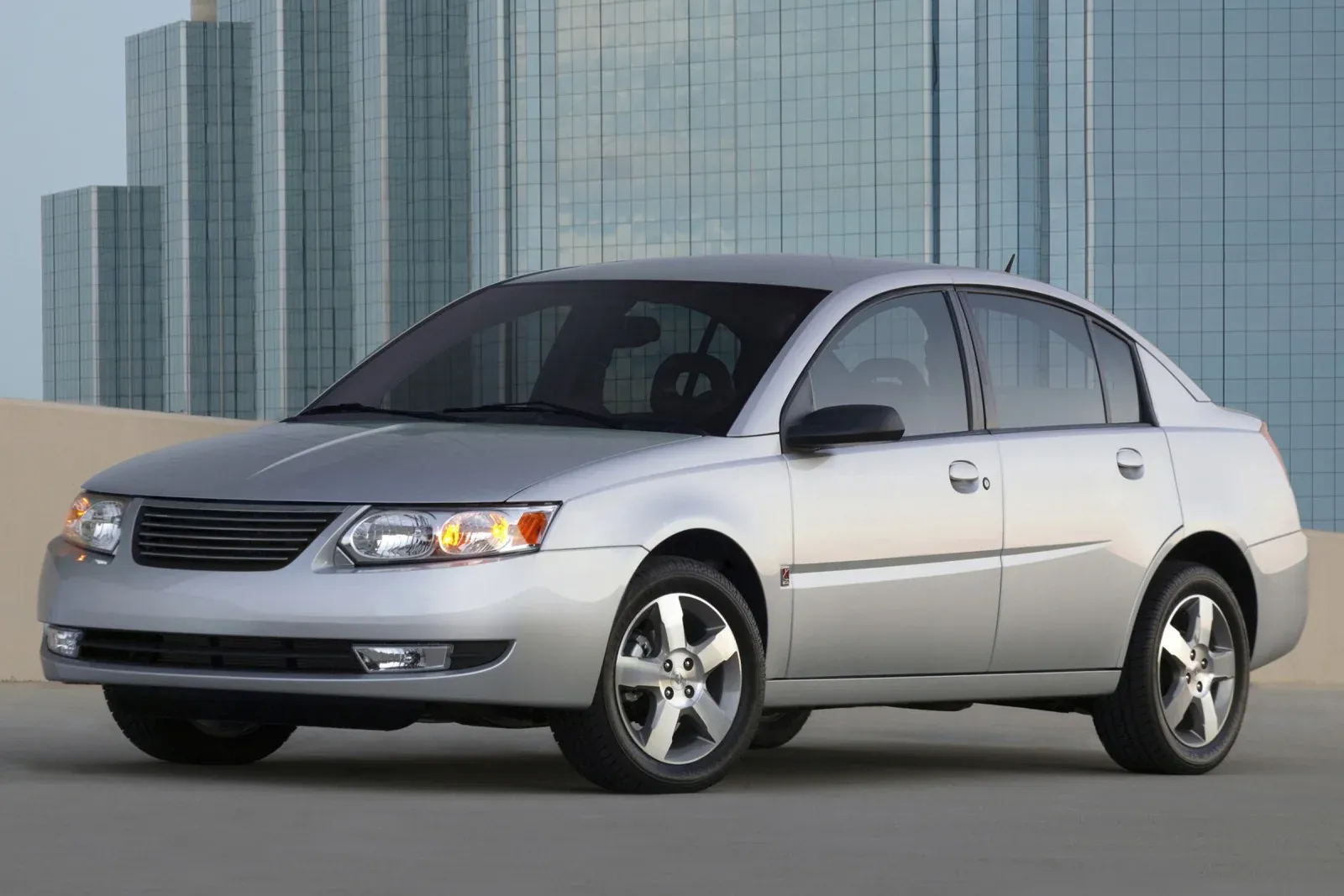 A silver Saturn Ion sedan is parked outdoors in front of a modern building with a glass facade.