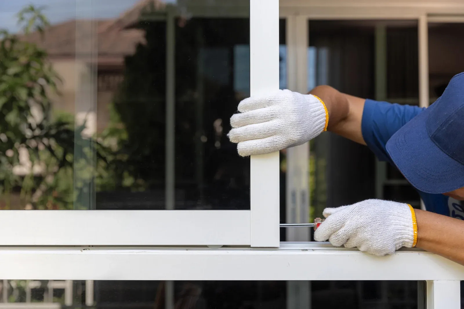 Window installer wearing gloves and a blue cap adjusts a white sliding window frame using a screwdri 