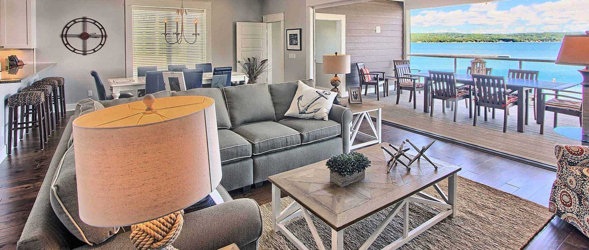 A spacious living area with a gray sofa, coffee table, and a large lamp in the foreground, with a dining area and an outdoor patio with a lake view in the background. The decor has a nautical theme, with an anchor pillow on the sofa.