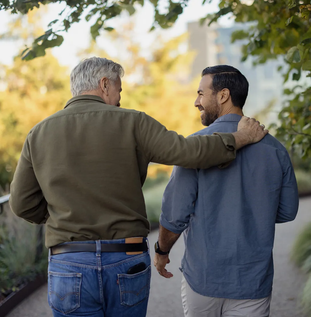 Two people standing outdoors with arms around each other's shoulders, smiling, against a backdrop of lush green trees.