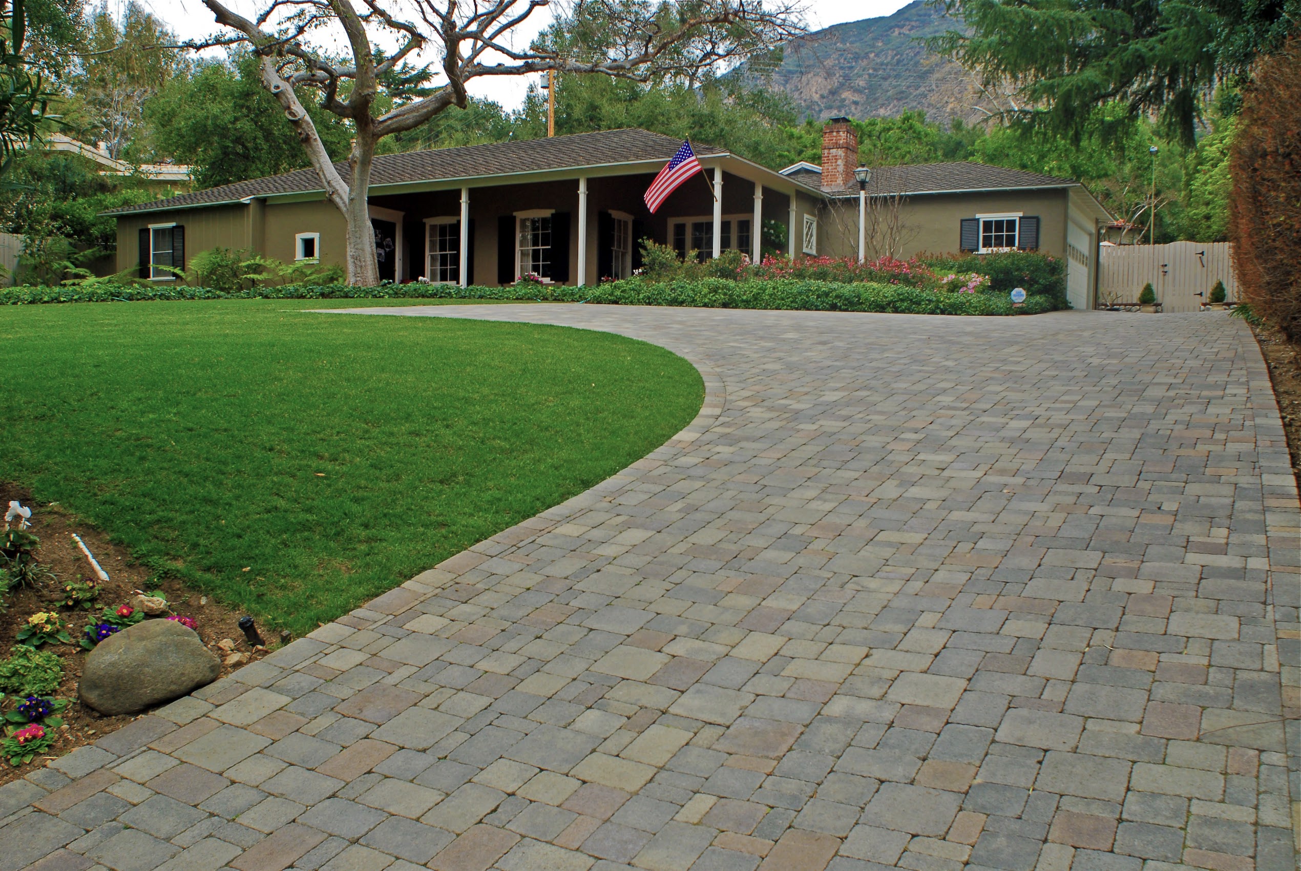Single-story home with an American flag, curved paver driveway, green lawn, mature trees, and mountain backdrop; peaceful, welcoming setting.