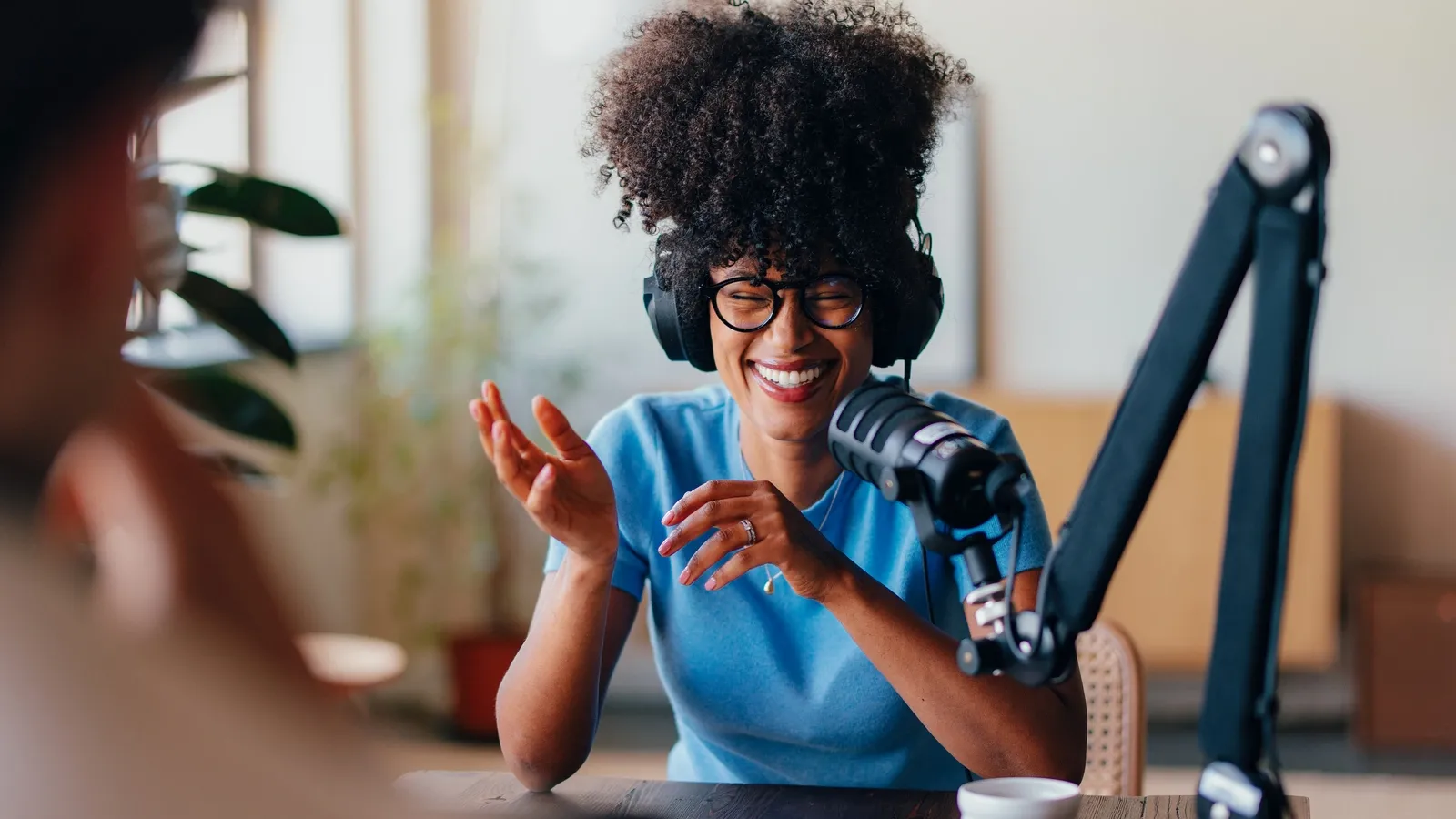 A woman with an afro and glasses, wearing headphones and a blue t-shirt, laughs while speaking into a microphone on a broadcast arm. She is seated at a wooden table in front of a blurred background that includes plants and windows.