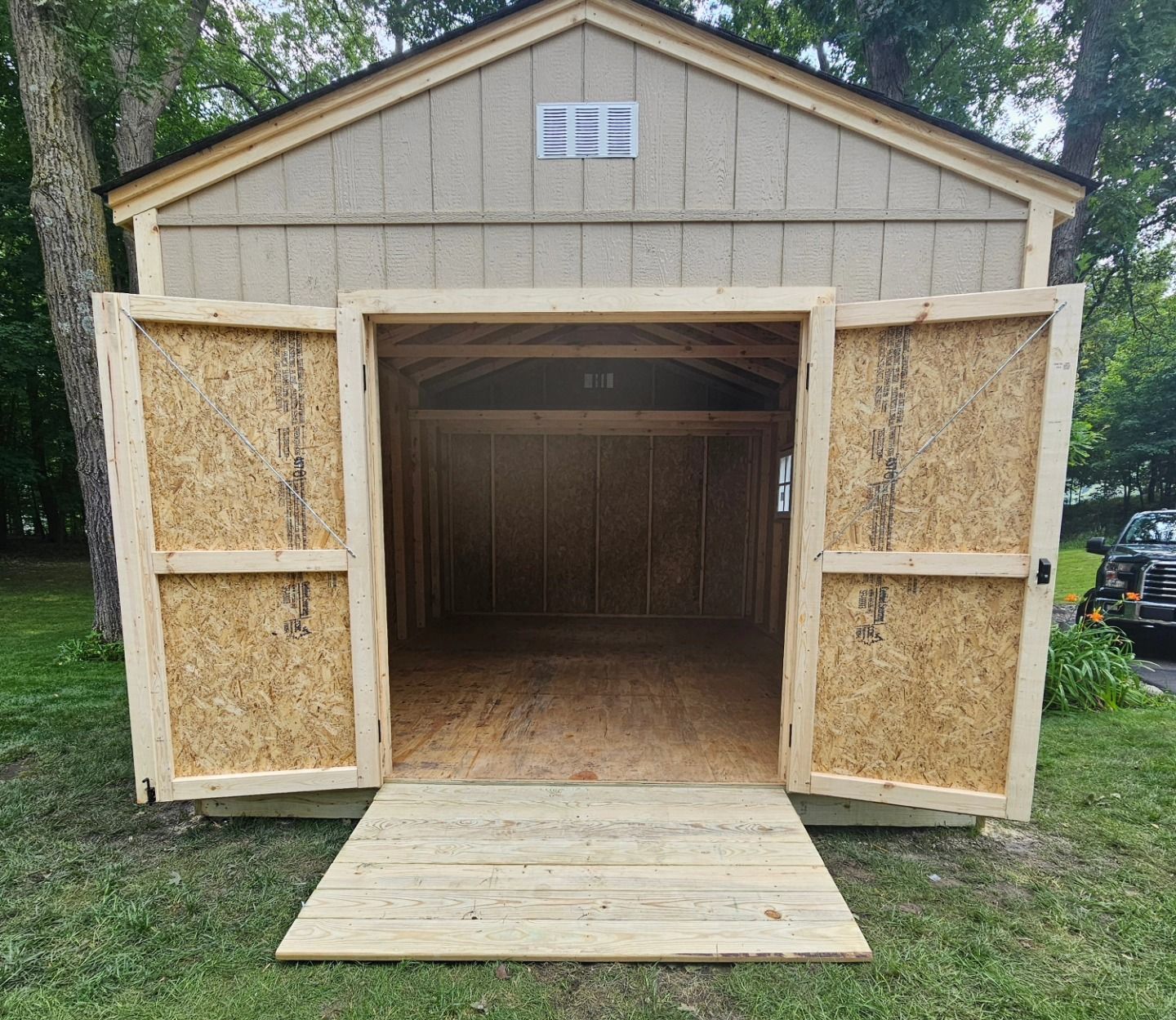 A newly built, tan-sided storage shed with a gable roof and wide, open double doors. A wooden ramp leads into the empty interior, which has visible wood framing and OSB wall sheathing.