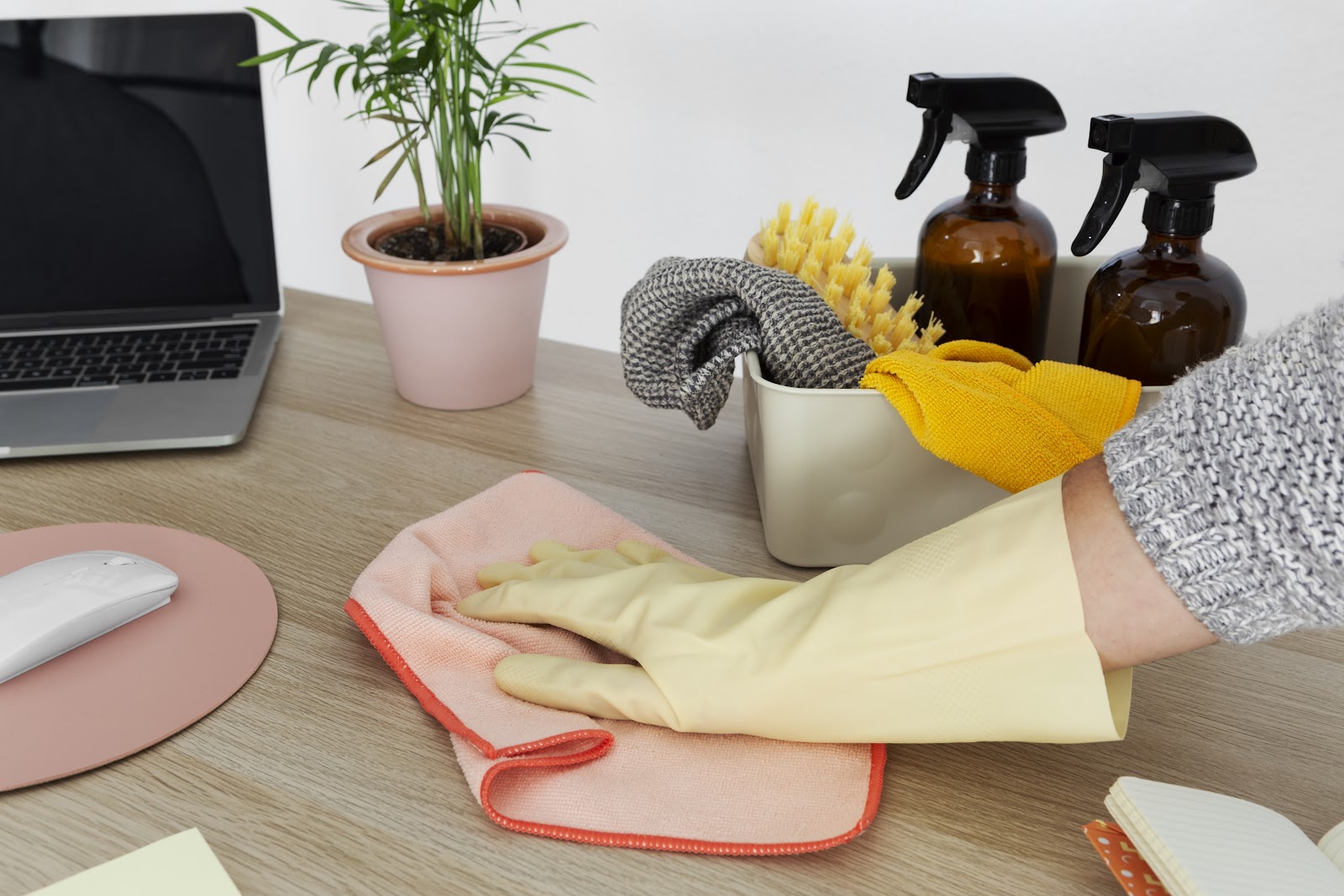 A gloved hand is cleaning a desk surface with a pink microfiber cloth. On the desk, there is a laptop, a mouse on a pink mat, a potted plant, a cleaning caddy filled with cleaning supplies including brushes and cloths, and two amber spray bottles. A person wearing a gray sweater is visible at the edge of the frame.