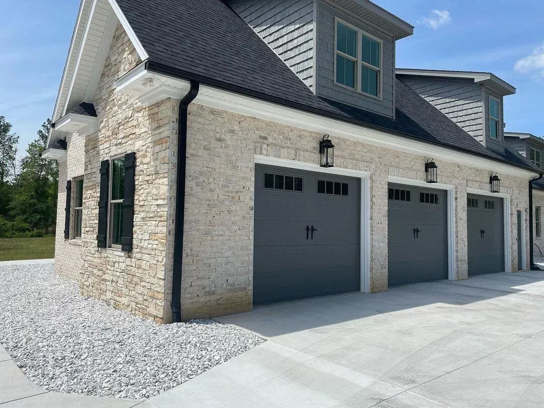 A large stone exterior features three matching gray sectional garage doors with small upper windows, dark hardware, lantern lights, and expansive concrete driveways surrounding the home with cohesive styling today.
