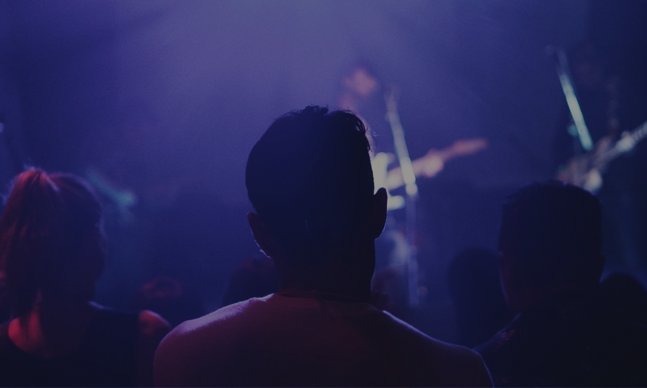 The back of three people's heads are visible in silhouette as they watch a band perform on a stage, illuminated by purple and blue stage lights. The band members are also silhouetted against the lighting, with guitars visible. The overall mood is dark and atmospheric, suggesting a live music event.