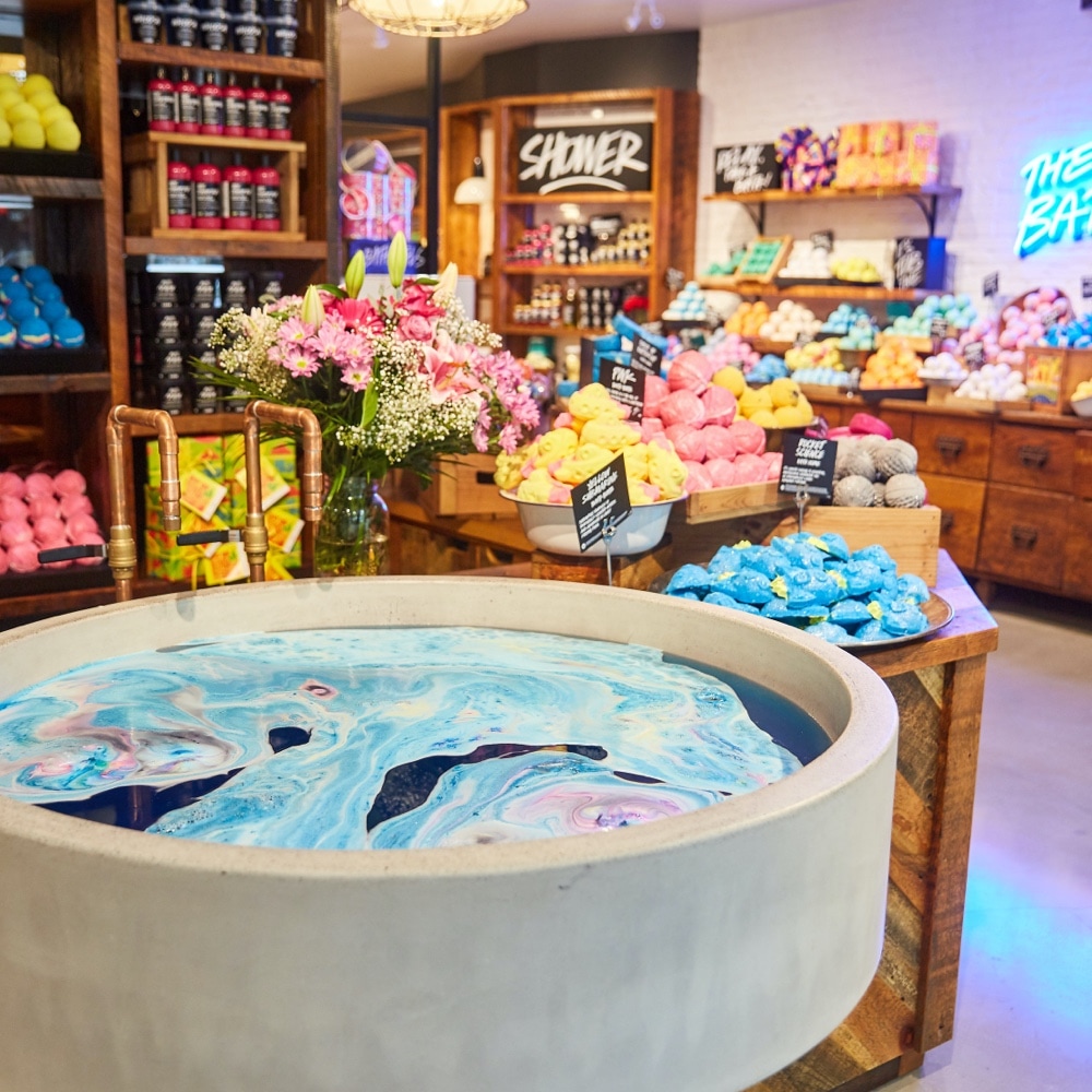 Inside a LUSH North America store where piles of bath bombs sit on a wooden table, organised by colour. Against the wall are colourful products and in the foreground is a large stone tub swirling with colourful, foaming water.