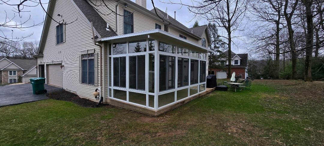 Spacious screened-in porch extension attached to a two-story home, featuring white framing, large mesh panels, and a flat roof. The structure enhances outdoor living, providing a protected area for relaxation while maintaining clear views of the backyard.