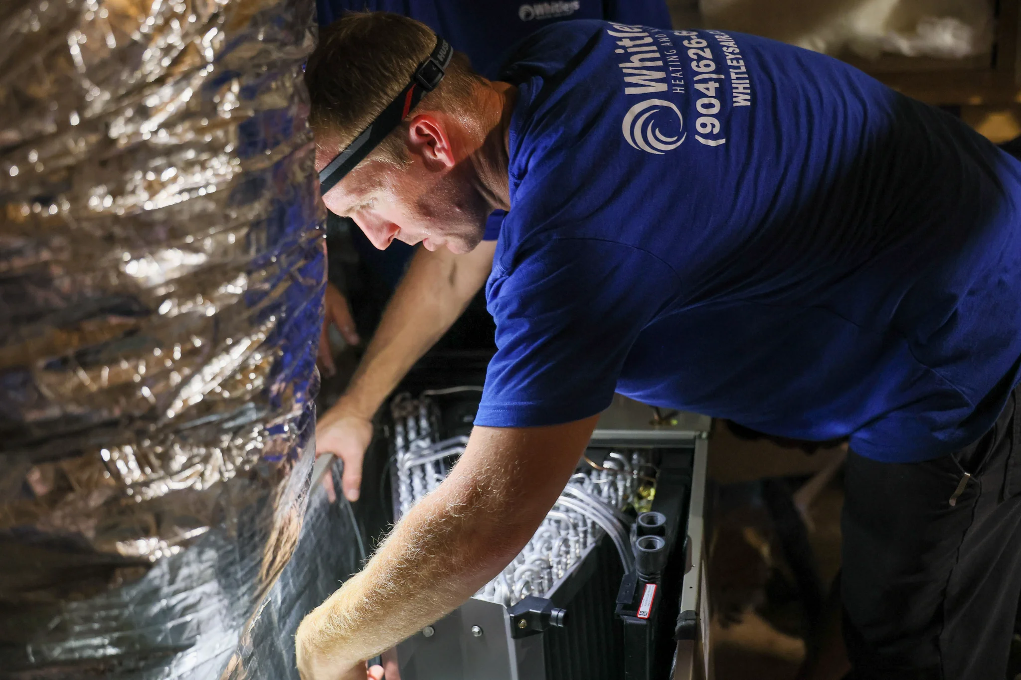 Two HVAC technicians are working on a cooling unit in a confined space with reflective insulation. O 