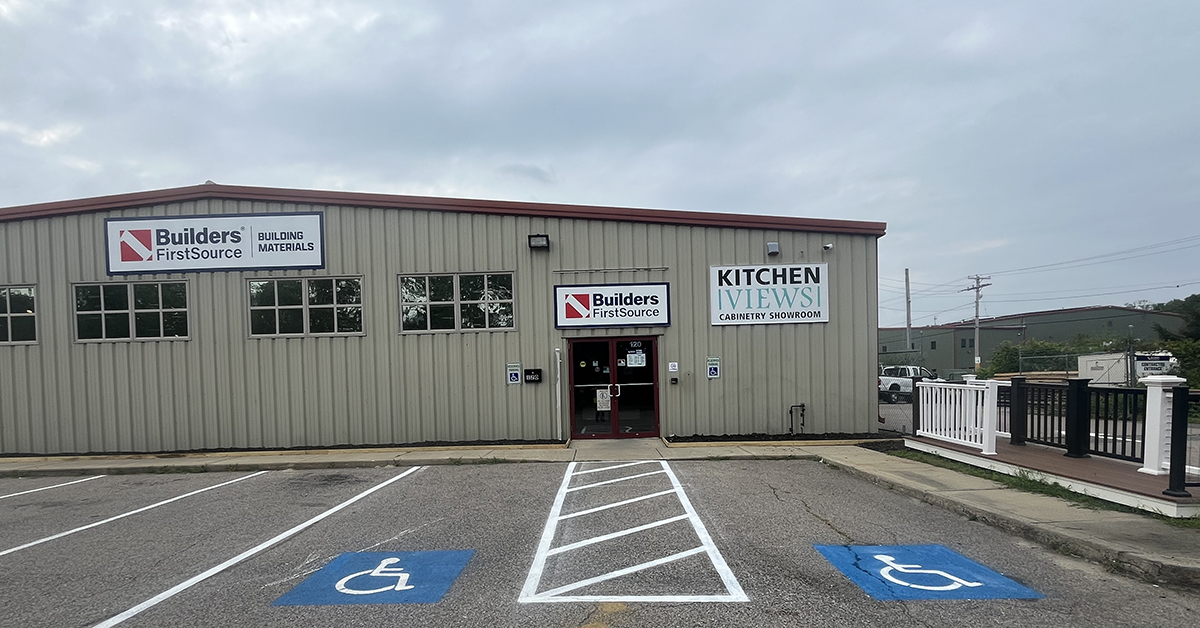 Exterior view of a Builders FirstSource Building Materials store and Kitchen Views cabinetry showroom, showing the storefront entrance, accessible parking spaces, and a composite deck railing display to the right of the building.