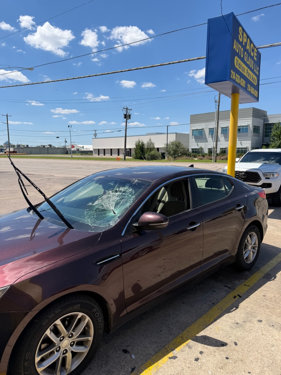 A maroon sedan with extensive windshield damage parked beneath the Space Auto Glass sign. The shattered glass shows when full windshield replacement may be necessary instead of a minor repair.