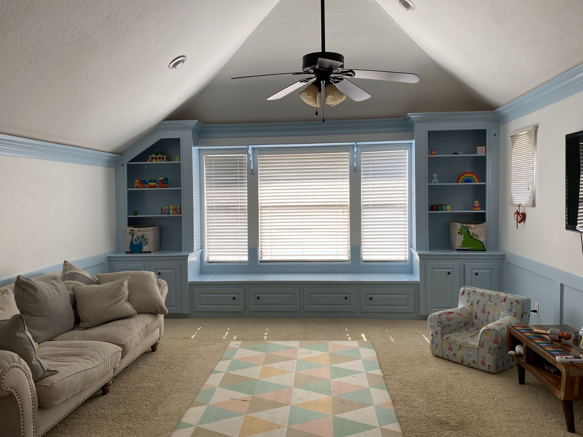 Bright living room featuring custom built-in shelving and window seating painted soft blue, complemented by vaulted ceilings, white walls, natural lighting, and a comfortable family-friendly atmosphere.