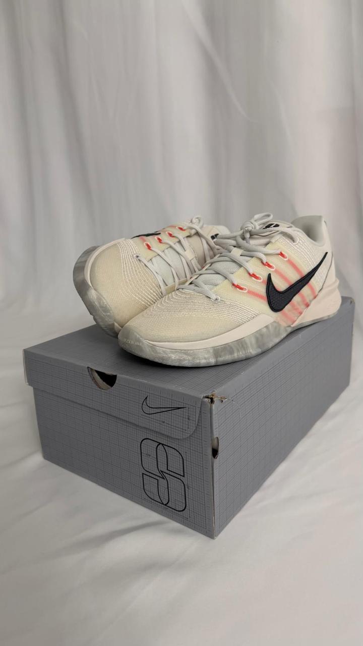 A pair of white Nike basketball shoes are displayed on top of a grey shoebox. The shoes feature a white knitted upper with subtle red accents and a black Nike swoosh. The shoebox has a grid pattern and the Nike logo.