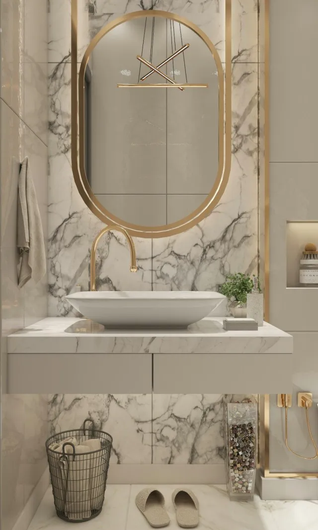 A modern bathroom vanity area featuring a large oval mirror with a gold frame, a white vessel sink with a gold faucet, a marble countertop and backsplash, a basket of towels, a pair of slippers, and a decorative glass container filled with small cubes. A contemporary pendant light hangs from the ceiling.