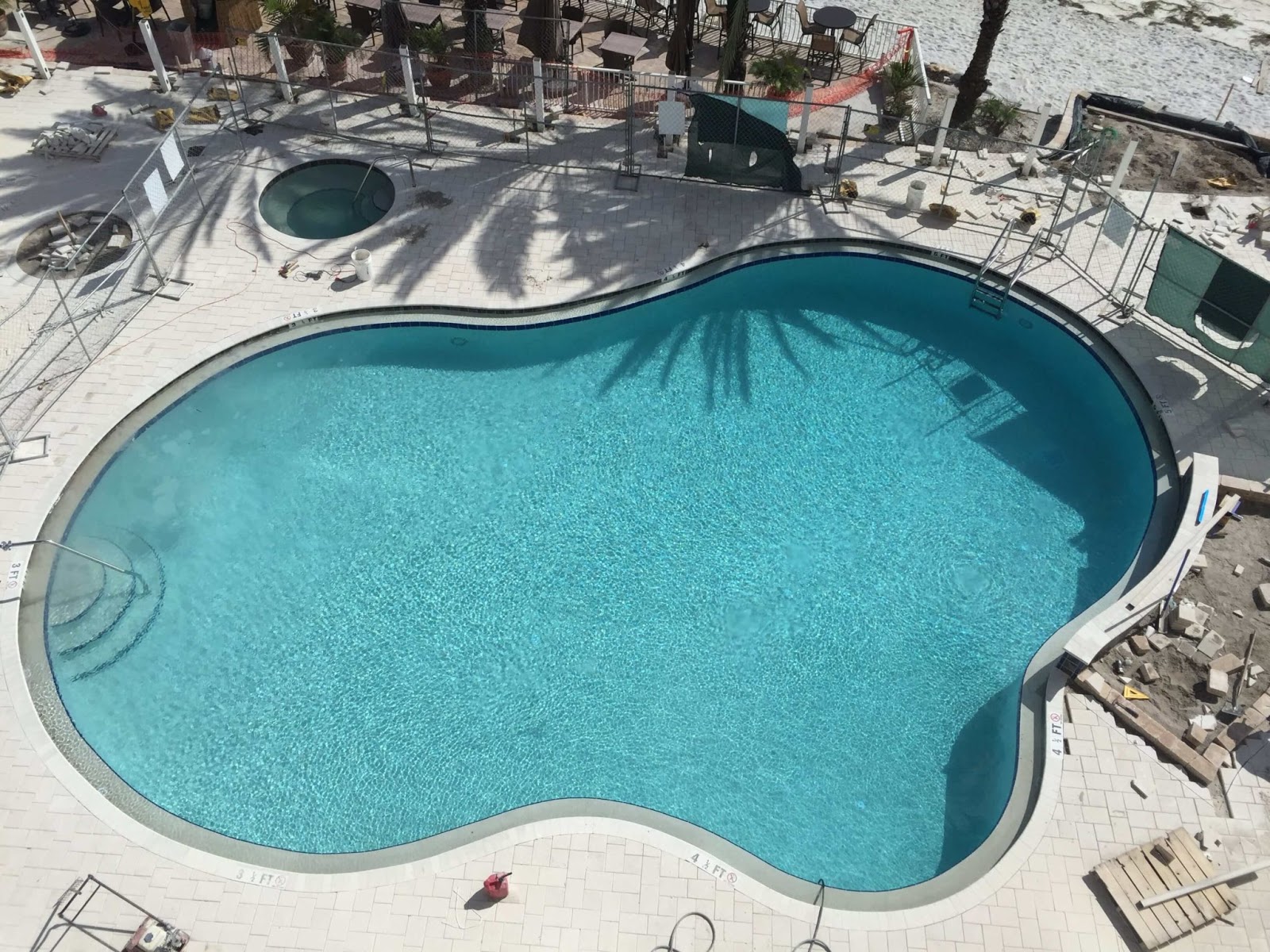 A custom residential swimming pool with clear blue water and built-in entry steps, surrounded by a light-colored paver patio, lush green lawn, and backyard landscaping.