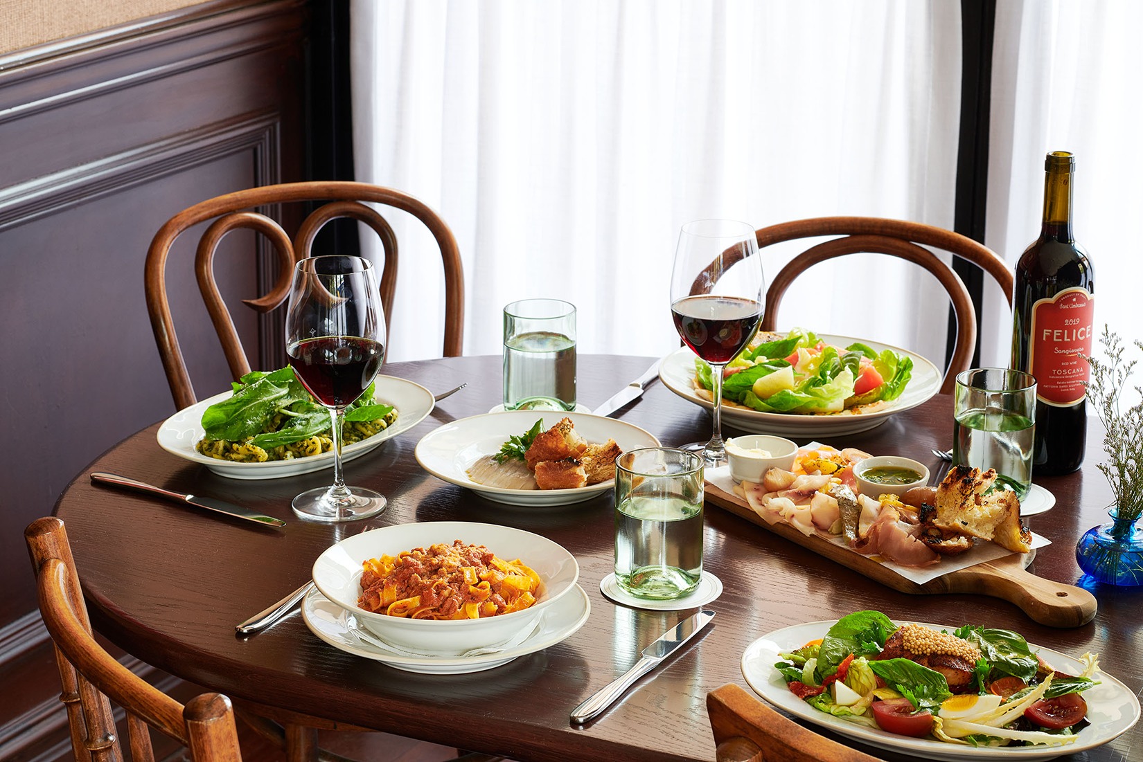 A round wooden table holds plates of pasta, salad, and a charcuterie board. Two glasses of red wine and a bottle of Felice Sangiovese are ready for a meal.