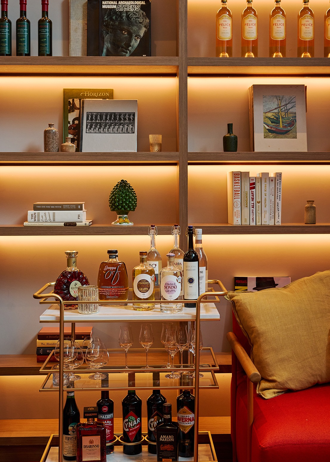 A sophisticated brass and marble bar cart featuring Grappa, Amaro, and Fernet-Branca. In the background, illuminated shelves display art books and Italian specialty bottles.