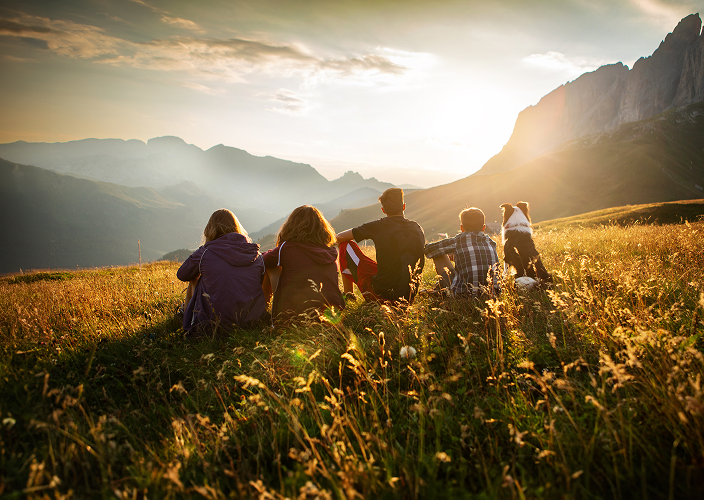 Un groupe d'amis et un chien assis dans un champ qui regardent le lever du soleil sur une chaîne de montagnes.