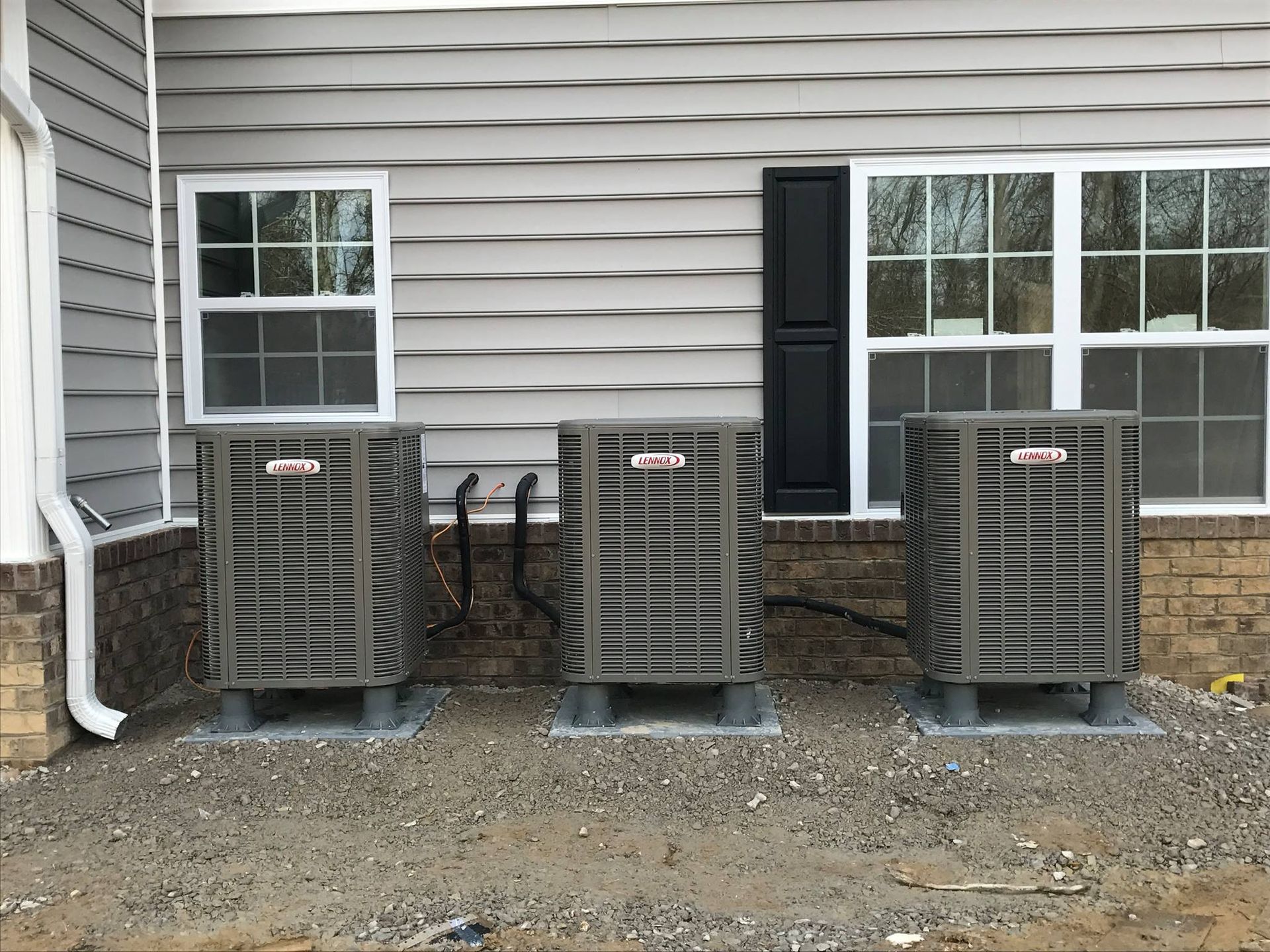 Three outdoor HVAC condenser units installed beside residential building, evenly spaced on concrete pads, connected with piping and wiring, showcasing new system setup for efficient cooling and professional installation work.