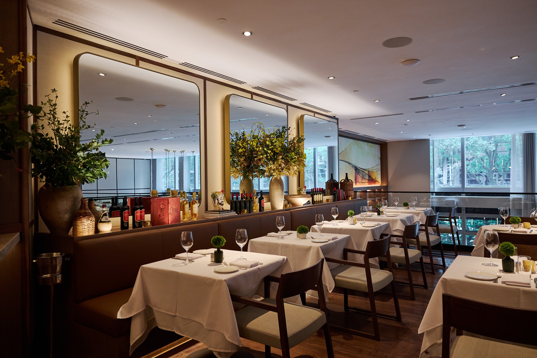 The main dining area of an Italian restaurant featuring brown leather banquettes, crisp white linens, and large mirrors reflecting a bright, airy atmosphere.