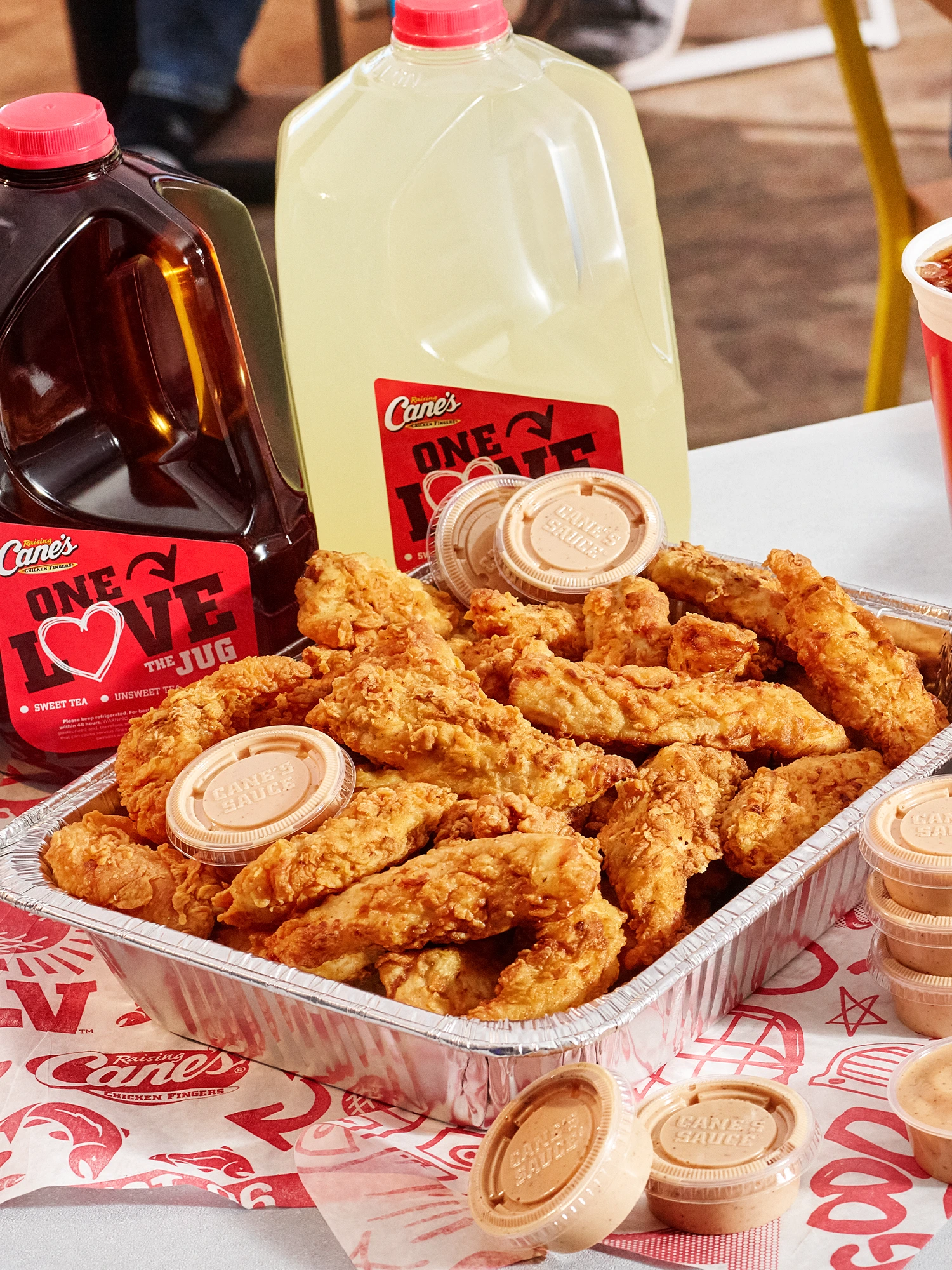 A large aluminum catering tray piled high with golden fried chicken fingers and several cups of Cane's sauce, sitting in front of two gallon jugs containing sweet tea and lemonade.