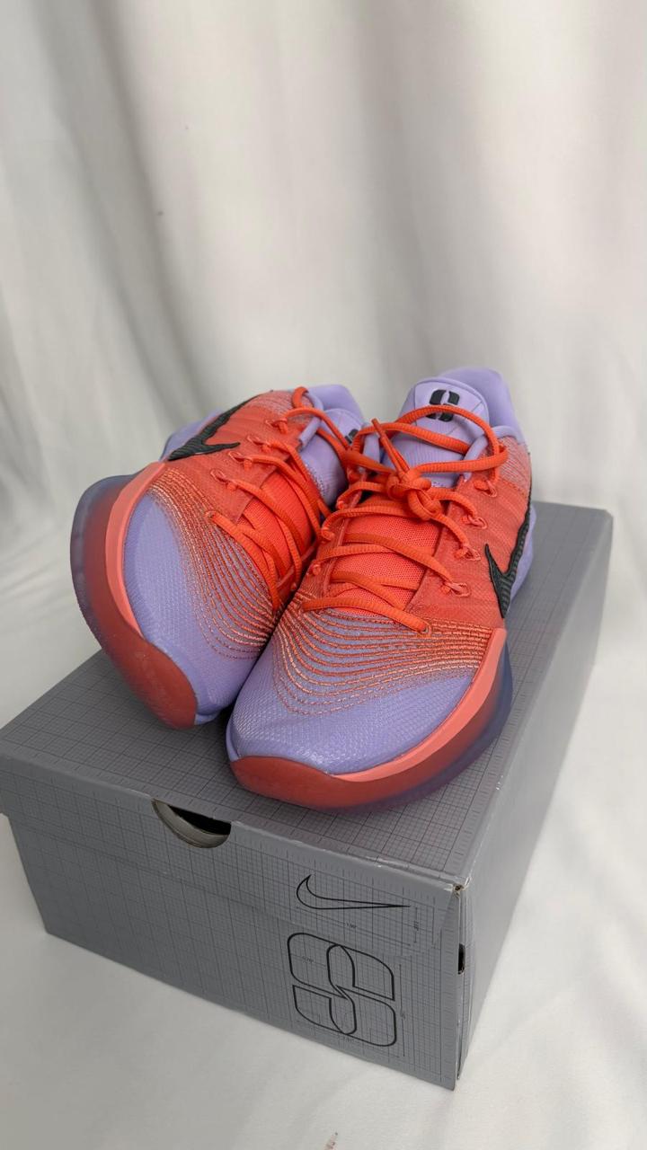 A pair of purple and orange Nike basketball shoes are displayed on a gray shoebox with a grid pattern and the Nike logo. The shoes feature black Nike swooshes on the sides and orange laces. The background is a plain white curtain.