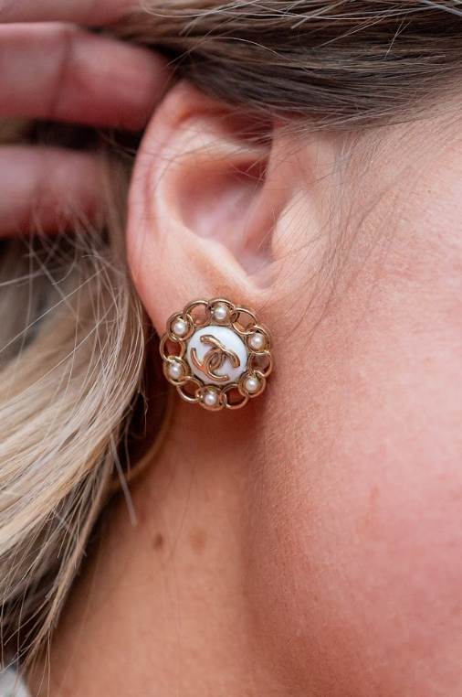A gold-toned floral earring features pearl-like accents, a white center, and an interlocking logo. Discover elegant statement earrings and curated jewelry at Eliana Curated in Old Town Alexandria, VA.