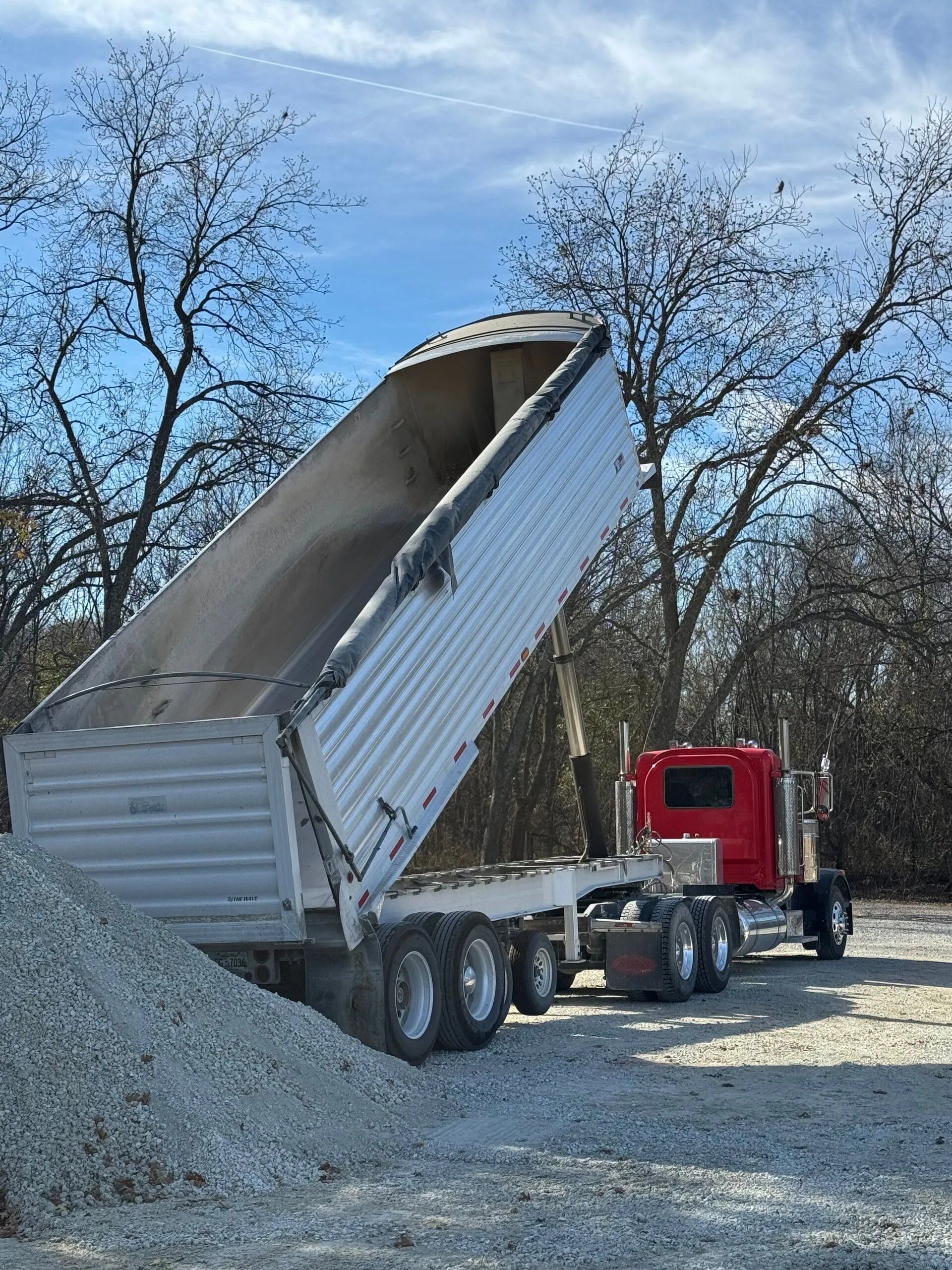 L & R Logistics LLC white and red dump truck raises its aluminum bed beside a pile of gravel, showing equipment used for aggregate hauling and delivery services for construction.
