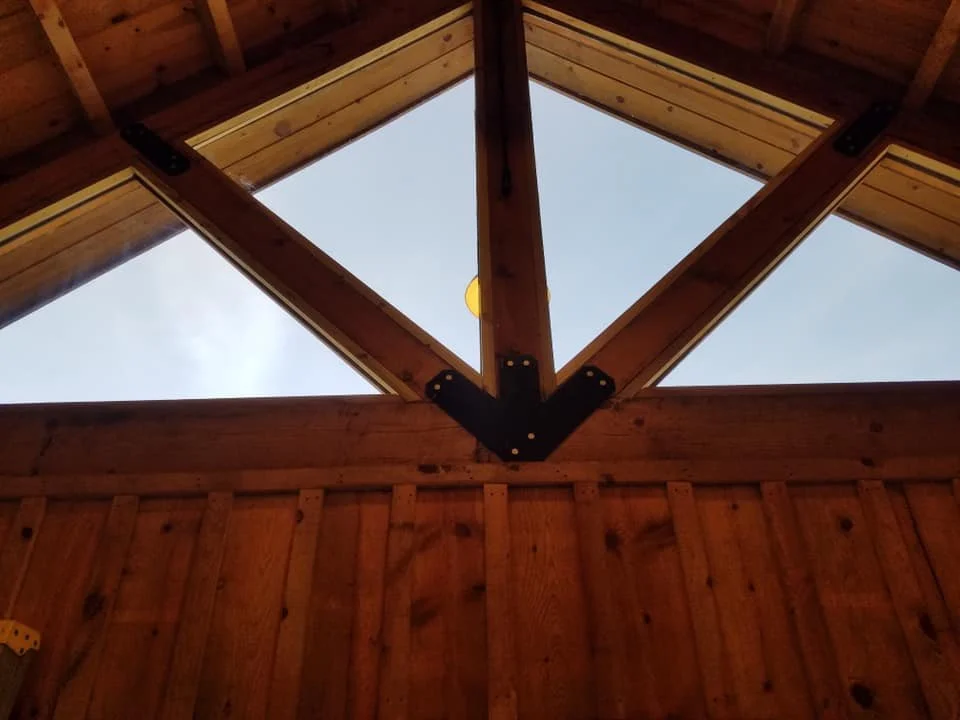 A view looking up into the triangular roof structure of a wooden building, with skylights revealing a light blue sky. Heavy wooden beams and rafters are visible, secured with black metal brackets. The interior walls are paneled with vertical wooden planks.