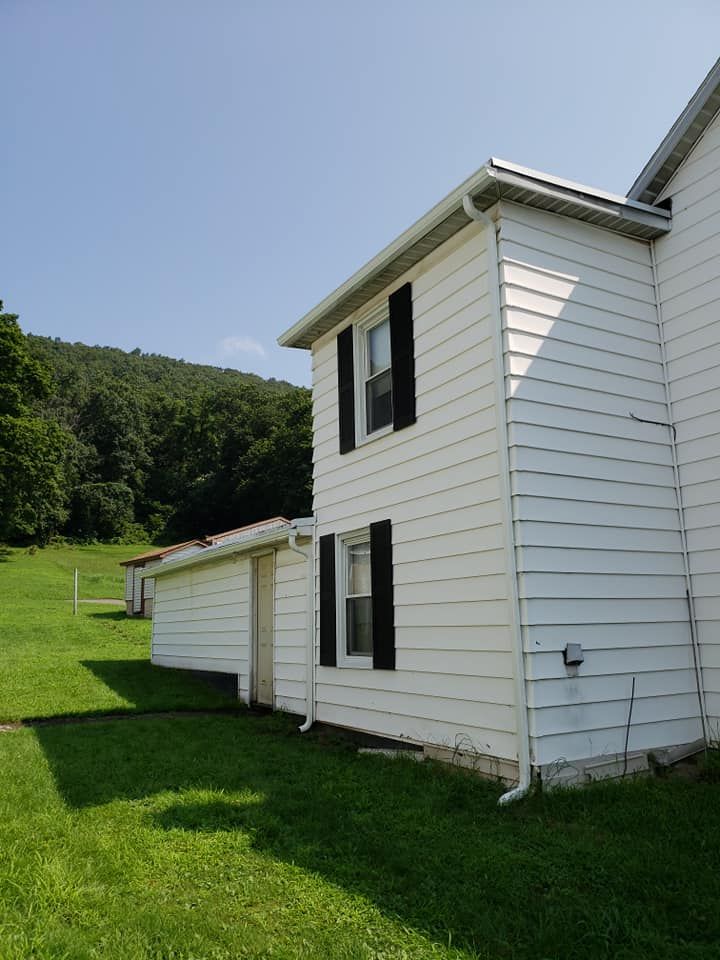 A white two-story house with horizontal siding and black shutters, featuring a small attached extension, set on a green lawn with trees and hills visible in the background.