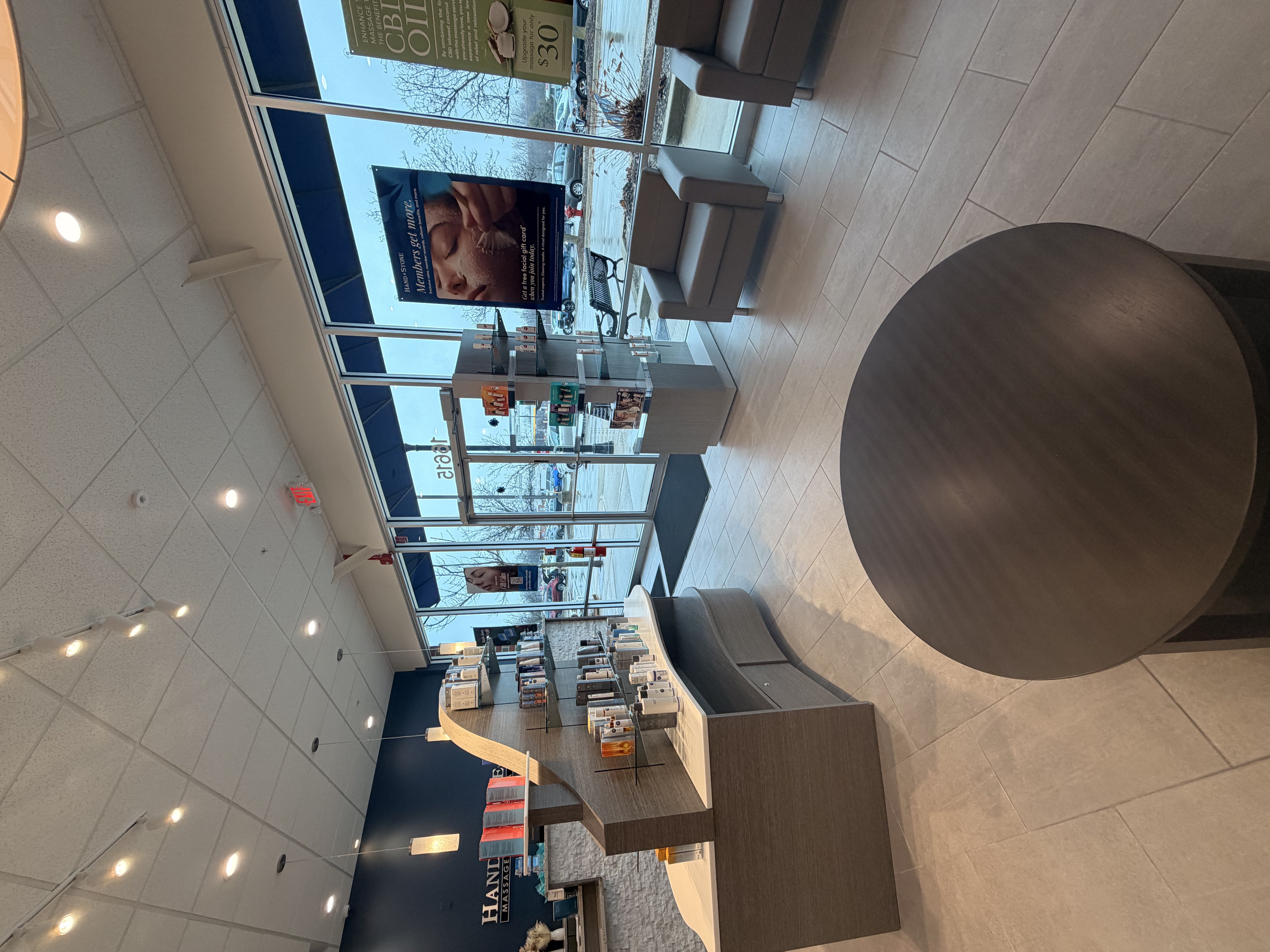 Reception area with curved desk and glass product shelving. Gray leather armchairs arranged near large windows. Round coffee table with dark wood finish. Navy blue wall with Hand & Stone branding visible in background. Natural light from storefront windows.