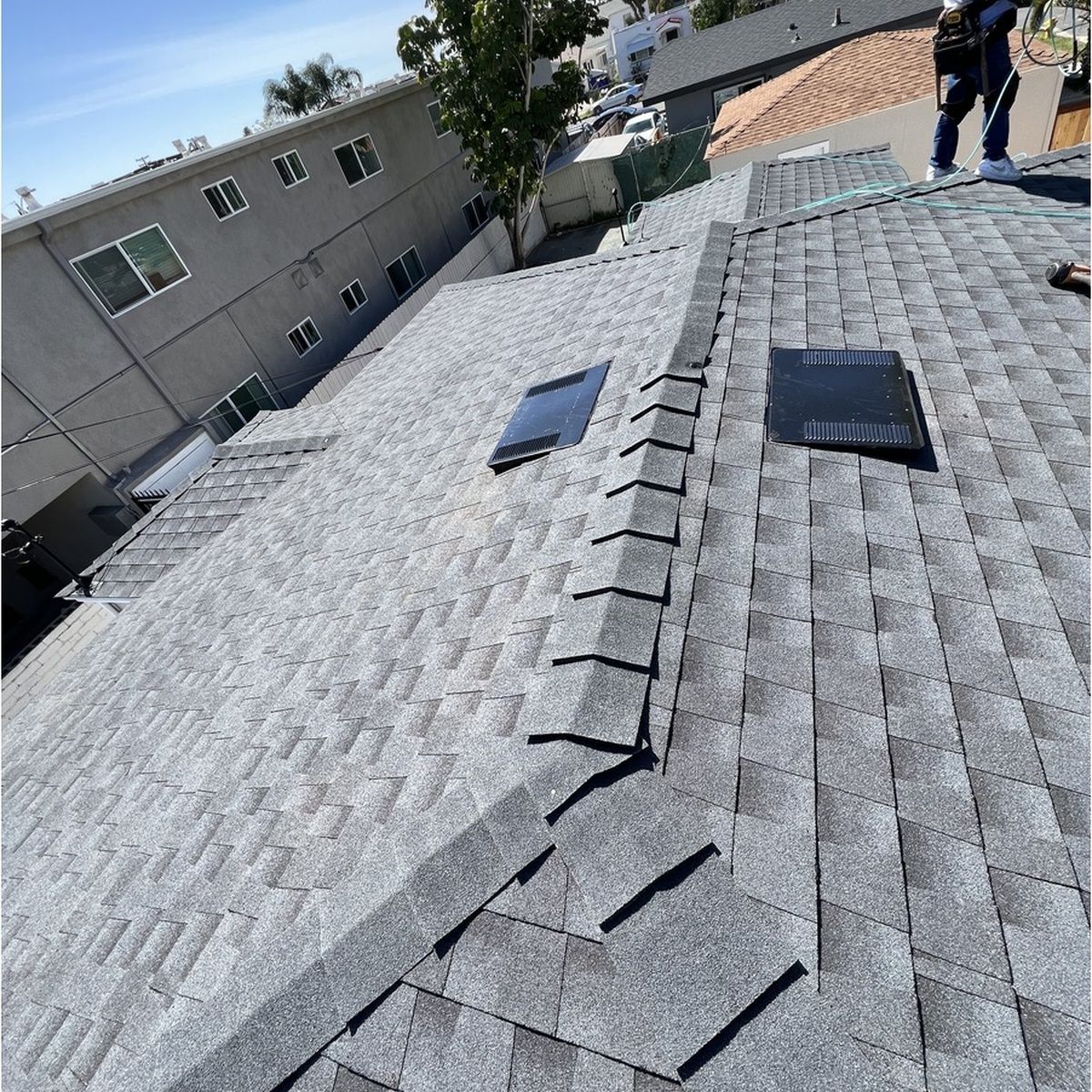 High-angle view showing a newly installed gray asphalt shingle roof with black roof vents, detailed ridge cap shingles, and a worker standing at the top against a residential backdrop.