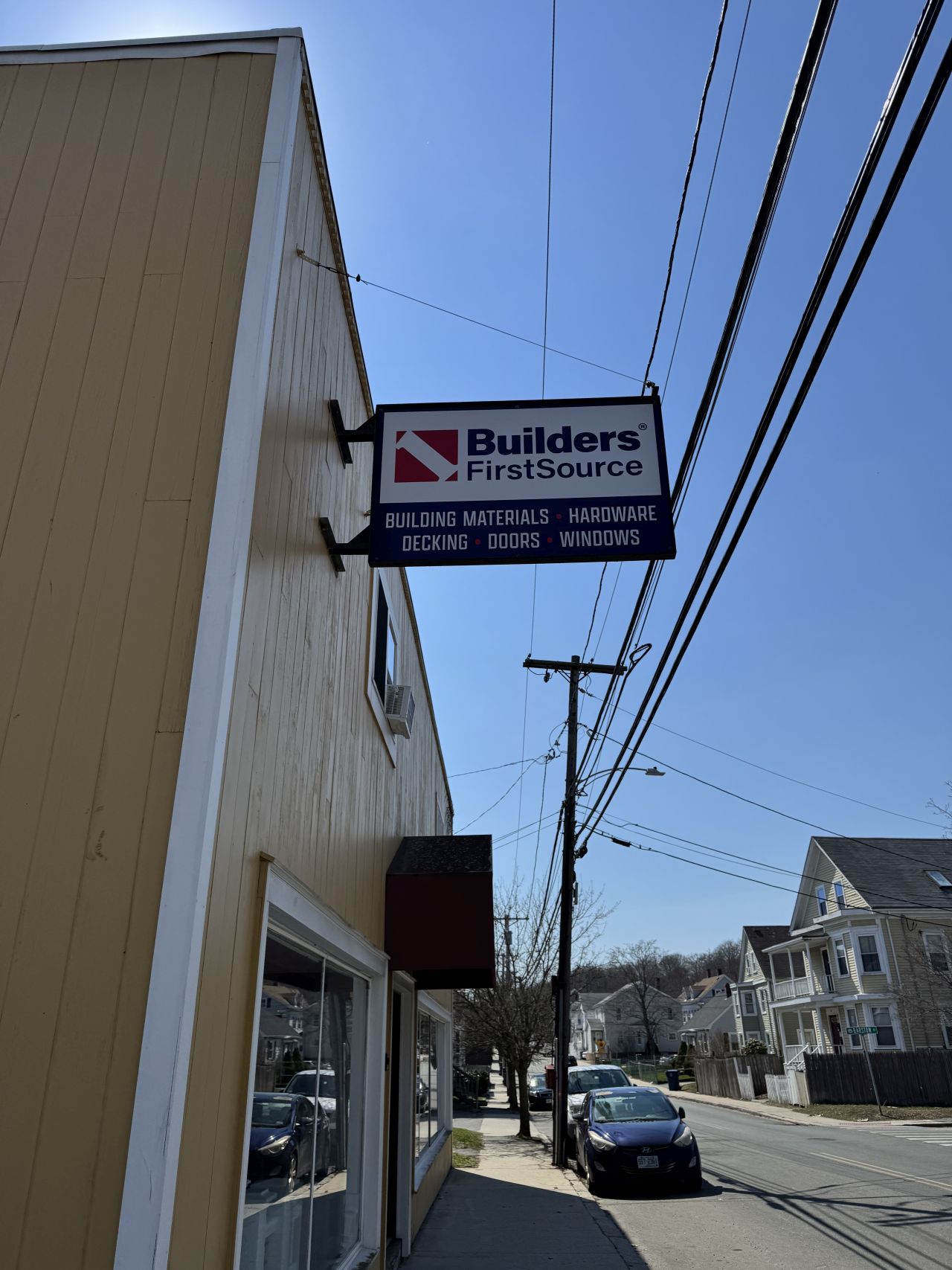 Hanging Builders FirstSource exterior sign mounted on a storefront, listing building materials, hardware, decking, doors, and windows.