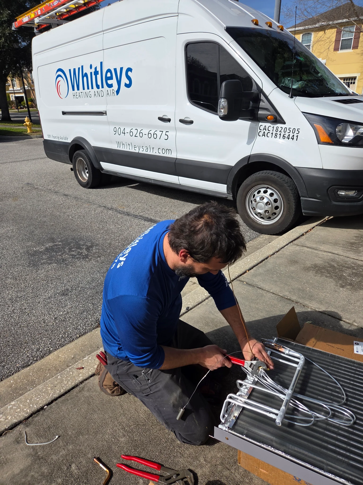 A technician wearing a blue shirt and dark pants is kneeling on the ground, working on an air condit 