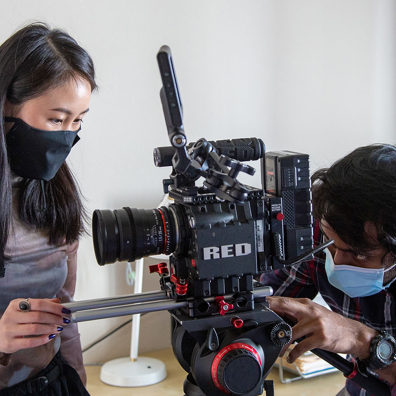 Two individuals, a woman and a man, wearing face masks, are operating a professional film camera mounted on a tripod. The woman on the left is wearing a black mask and a greyish top, and she is looking down at the camera with her left hand resting on a silver rail. The man on the right is wearing a light blue face mask and a plaid shirt. He is adjusting the camera with his right hand. The camera is a RED brand camera, identifiable by the prominent red logo. The background is a plain, light-colored wall with some indistinct objects on the right.
