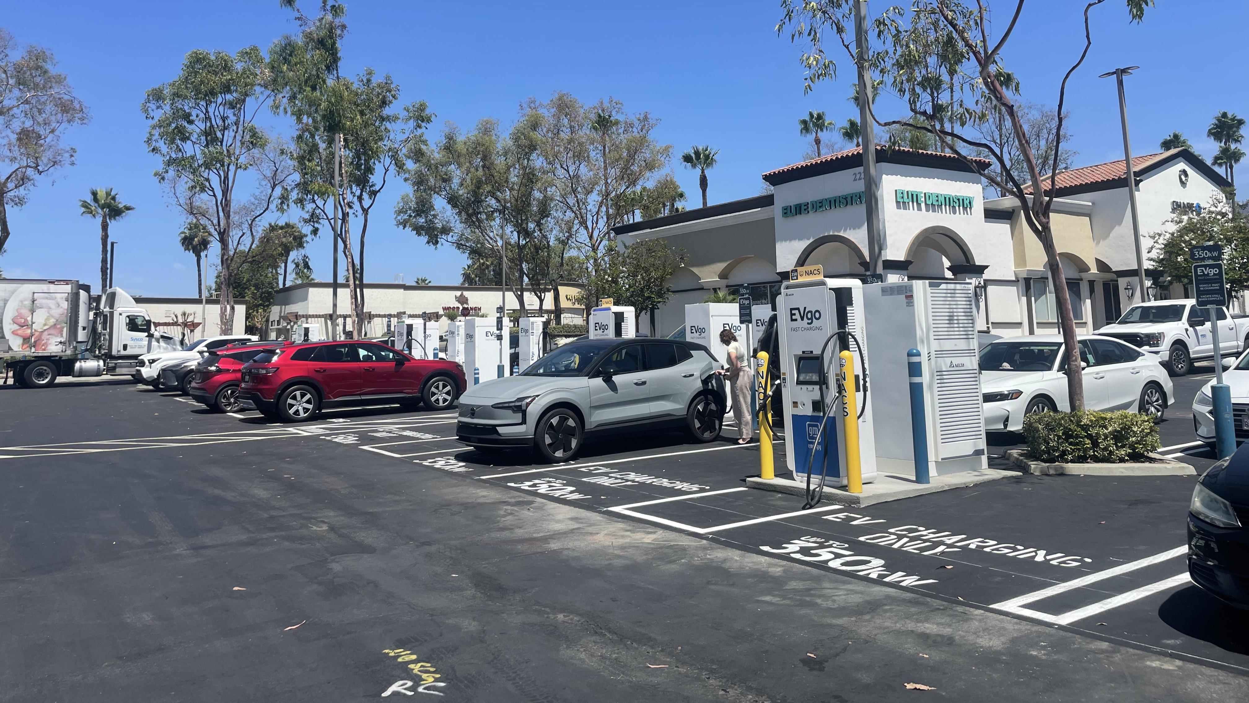 Several EVgo fast charging units installed in marked parking spaces at a public charging station, with EVgo logos on the chargers, protective bollards around each unit. NACS and CCS chargers featured in the image.