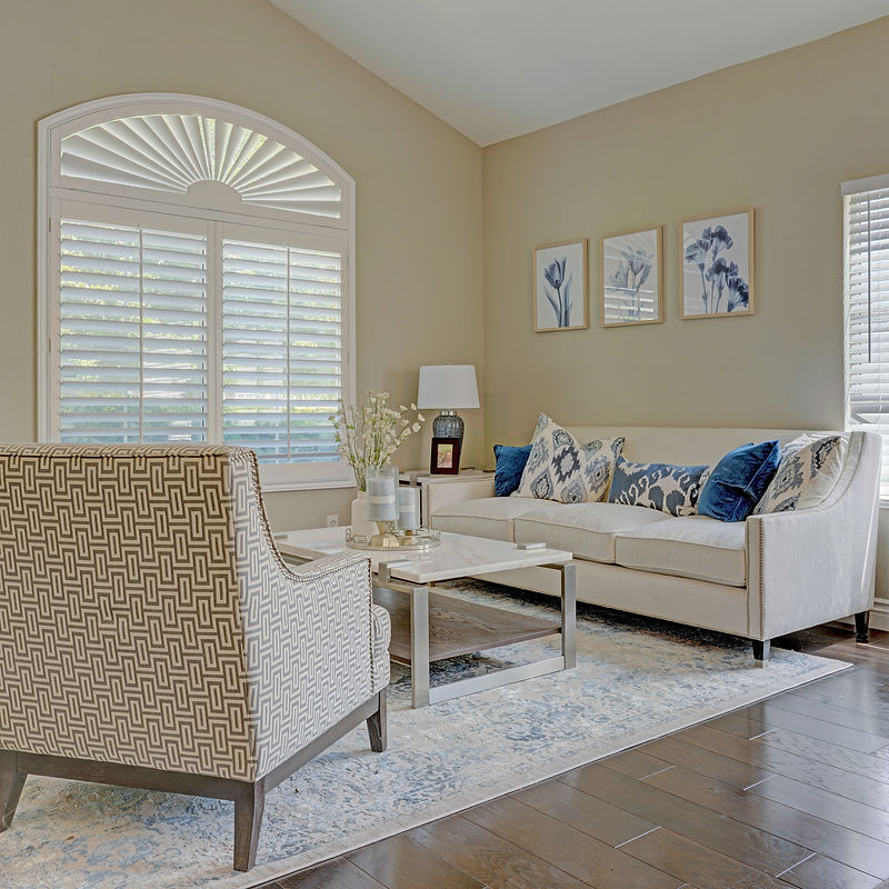 A well-decorated living room features a patterned armchair, a white sofa with blue and white patterned pillows, a marble-topped coffee table, and a large arched window with white shutters.