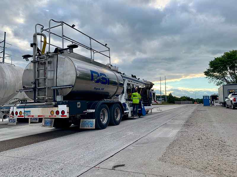 Low-angle shot under overcast skies showcasing a large, stainless steel DSI tanker trailer parked alongside a specialized diamond grinding rig on concrete pavement. The reflective chrome tank bears the DSI logo with work trailers and support vehicles parked off to the right.
