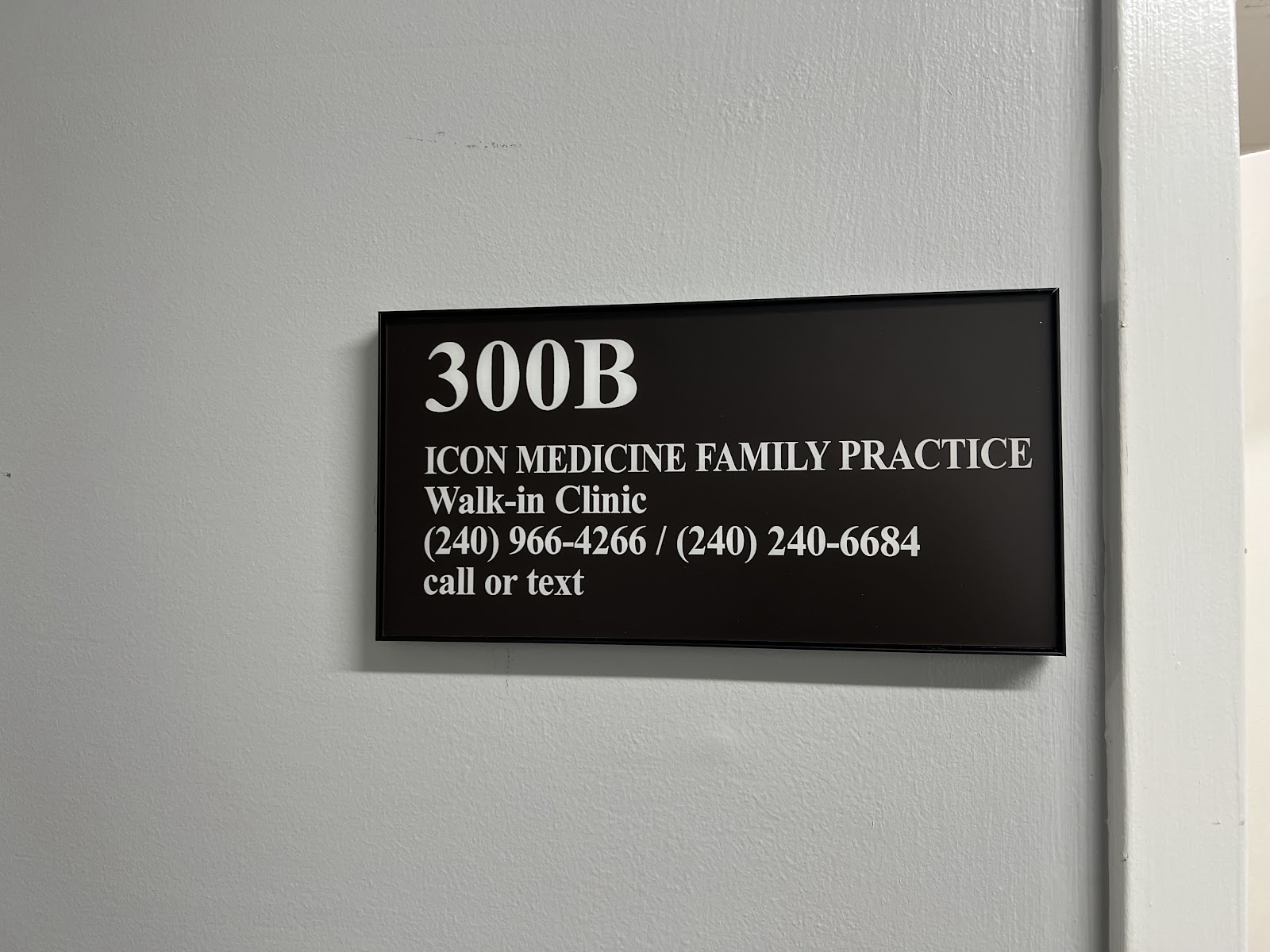 A black sign with white text is mounted on a light gray wall. The sign reads "300B", followed by "ICON MEDICINE FAMILY PRACTICE", "Walk-in Clinic", and then two phone numbers: "(240) 966-4266 / (240) 240-6684". Below the phone numbers, it says "call or text". The sign has a thin black frame.